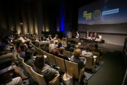 behind the audience in auditorium facing a stage with three people on it