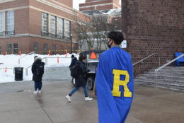 the back of a male student wearing blue cape and a yellow R and rush rhees library tower in background