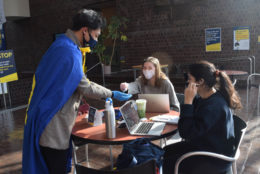 man in blue cape handing our i heart UR pins to two women sitting at table with their laptops