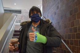 green casted wrist with thumbs up by a man wearing a hat and mask holding a coffee
