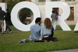 man and woman sitting on skateboard in quad in front of meliora letters
