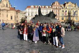 group of people posing in front of a monument in center of a plaza with buildings and blue sky in background