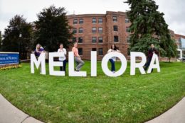 four women and a man posing with large white meliora letters
