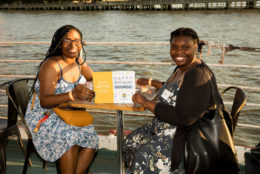 two women seated at table on a boat with water in background posing for photo