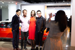 three men and a woman posing in front of classic car while woman taking photo with cell phone