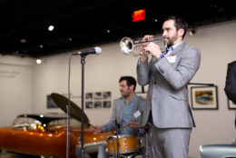 man in gray suit playing the trumpet with drummer in background