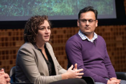 woman speaking on stage seated next to a man wearing purple sweater