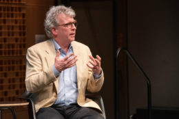 man in tan jacket and glasses speaking on stage