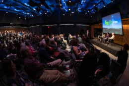 audience in stadium seating with four speaking seated on stage and large screen on stage