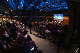 audience in stadium seating with four speaking seated on stage and large screen on stage