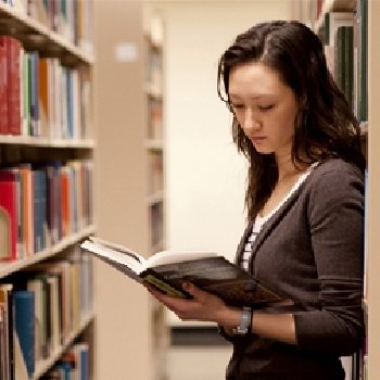 an woman is standing as she reads a book in an aisle within a library.