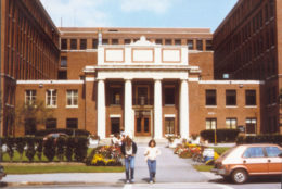 old photo of SMD entrance a brick building with four pillars and two people walking out across the street