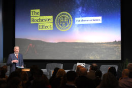 man on stage speaking to audience with large screen in background