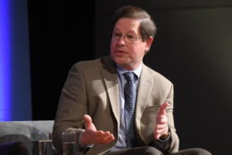 man seated in suit and tie on stage speaking