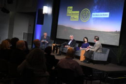 three men and woman on stage seated giving talk at event with large screen in background