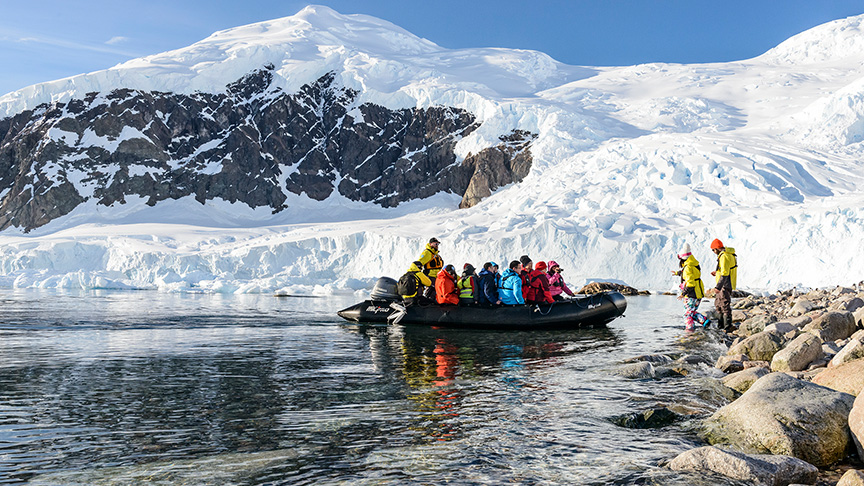 People on a raft boat near a rocky shore with ice capped mountains in the background