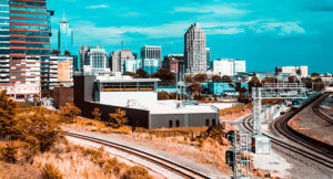 A view of downtown Raleigh, North Carolina, showing modern high-rise buildings, a railway line curving in the foreground, and a clear blue sky above.