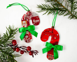 Two red circuit board ornaments shaped like candy canes, each decorated with a green ribbon bow and hanging by green strings, are displayed among evergreen branches and red berries.