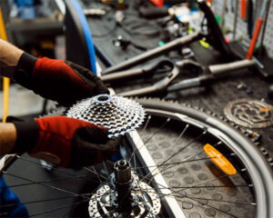 A person wearing red and black gloves is installing a bicycle gear cassette onto a wheel hub in a workshop with various bike tools and parts in the background.