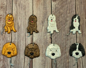 A set of eight handmade dog ornaments, each featuring different dog breeds in various colors with metallic finishes, displayed on a wooden background in two rows.