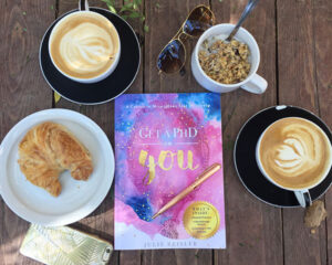 A flat lay image featuring the book Get a PhD in You by Julie Reisler surrounded by two lattes, a bowl of granola, a croissant, sunglasses, a pen, and a smartphone on a wooden table.