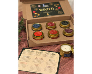 A boxed Winter Candle Collection set from Le Silo Bleu displayed on a wooden surface, containing six small gold-lidded candles with a descriptive card listing the included seasonal scents.