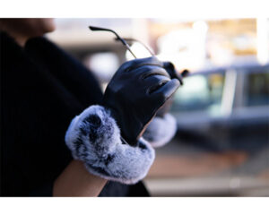 A close-up image of a person wearing black leather gloves with gray faux fur cuffs, holding a pair of sunglasses in an outdoor urban setting.