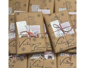 A display of wrapped books labeled “Victory Books,” each tied with twine and accompanied by small decorative cards and stickers on brown paper packaging.