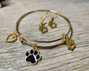 A gold bracelet and matching earrings featuring small heart and paw-print charms are displayed on a wooden surface.