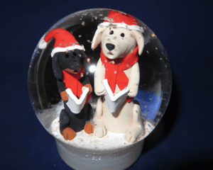 A glass snow globe contains two festive dog figurines wearing red hats and scarves, standing together on a snowy base.