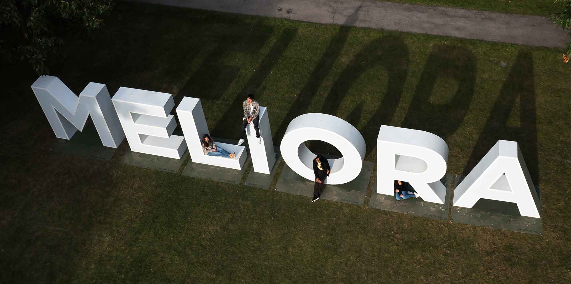Meliora letters from above on green lawn with people sitting on some of the letters
