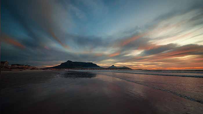 An evening sky with clouds, and a beach in the foreground with a small silhouette of a mountain in the background