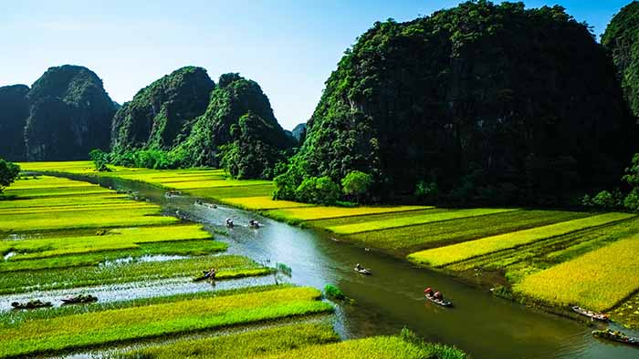 People canoeing on a river, with green fields on either sid. And green mounds of mountains in the background