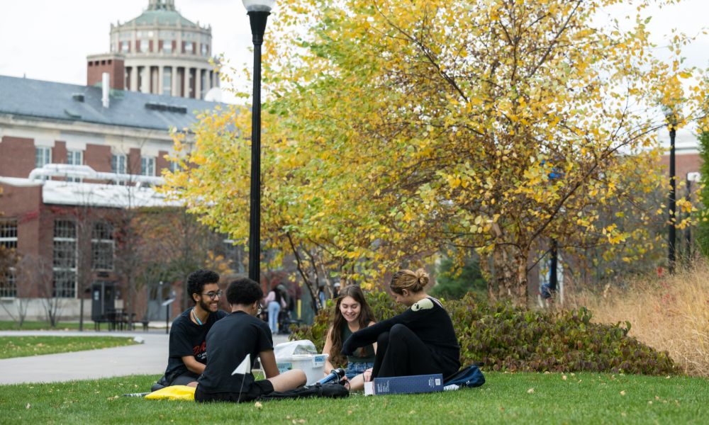 Students relax on the grass in front of the University of Rochester library, enjoying a sunny day outdoors.