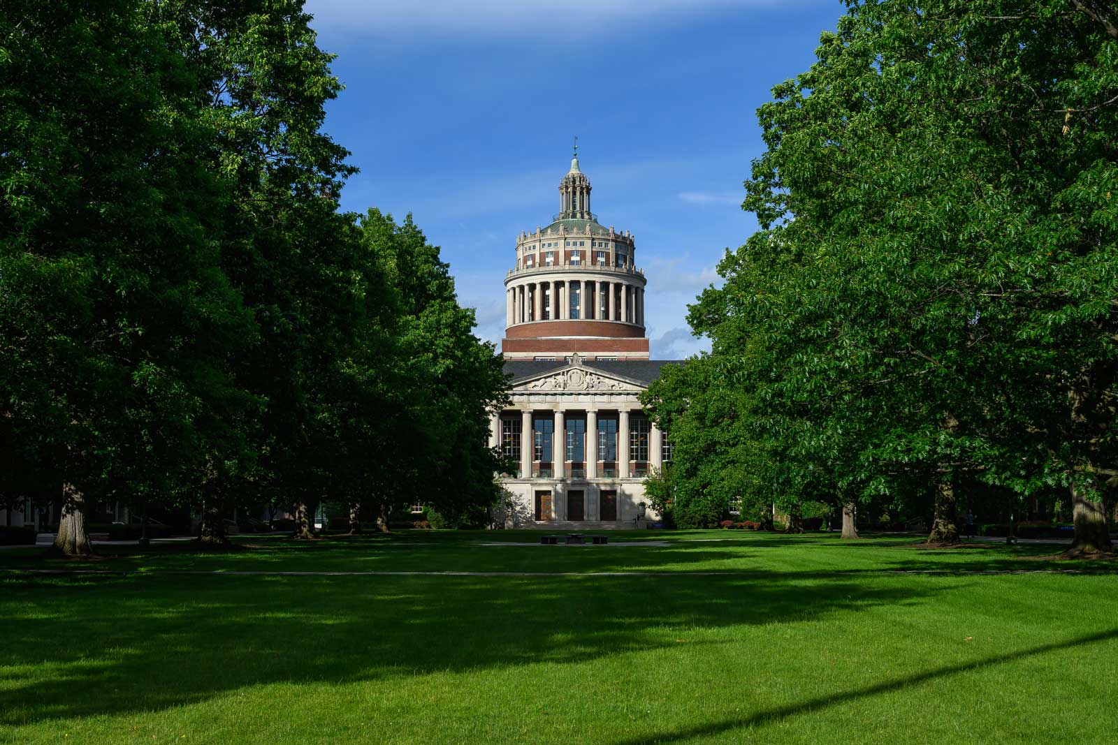 Rush Rhees Library stands at the end of a green campus quad, framed by tall leafy trees beneath a blue sky.