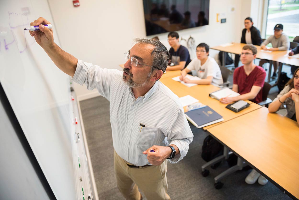 Professor writing on a whiteboard at the front of a classroom