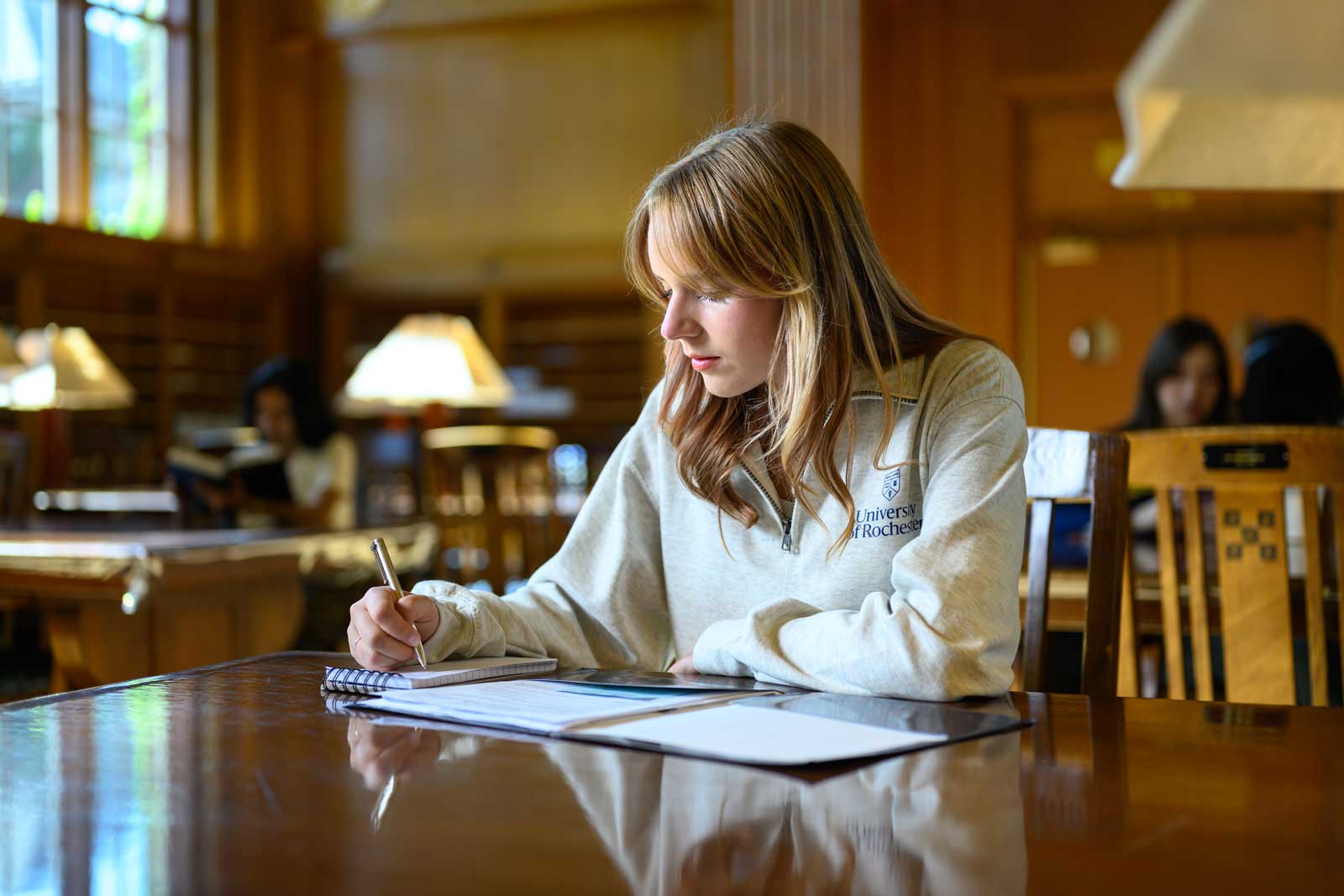 Window light falls across a student writing in a notebook, surrounded by warm wood and glowing lamps in a quiet library reading room.