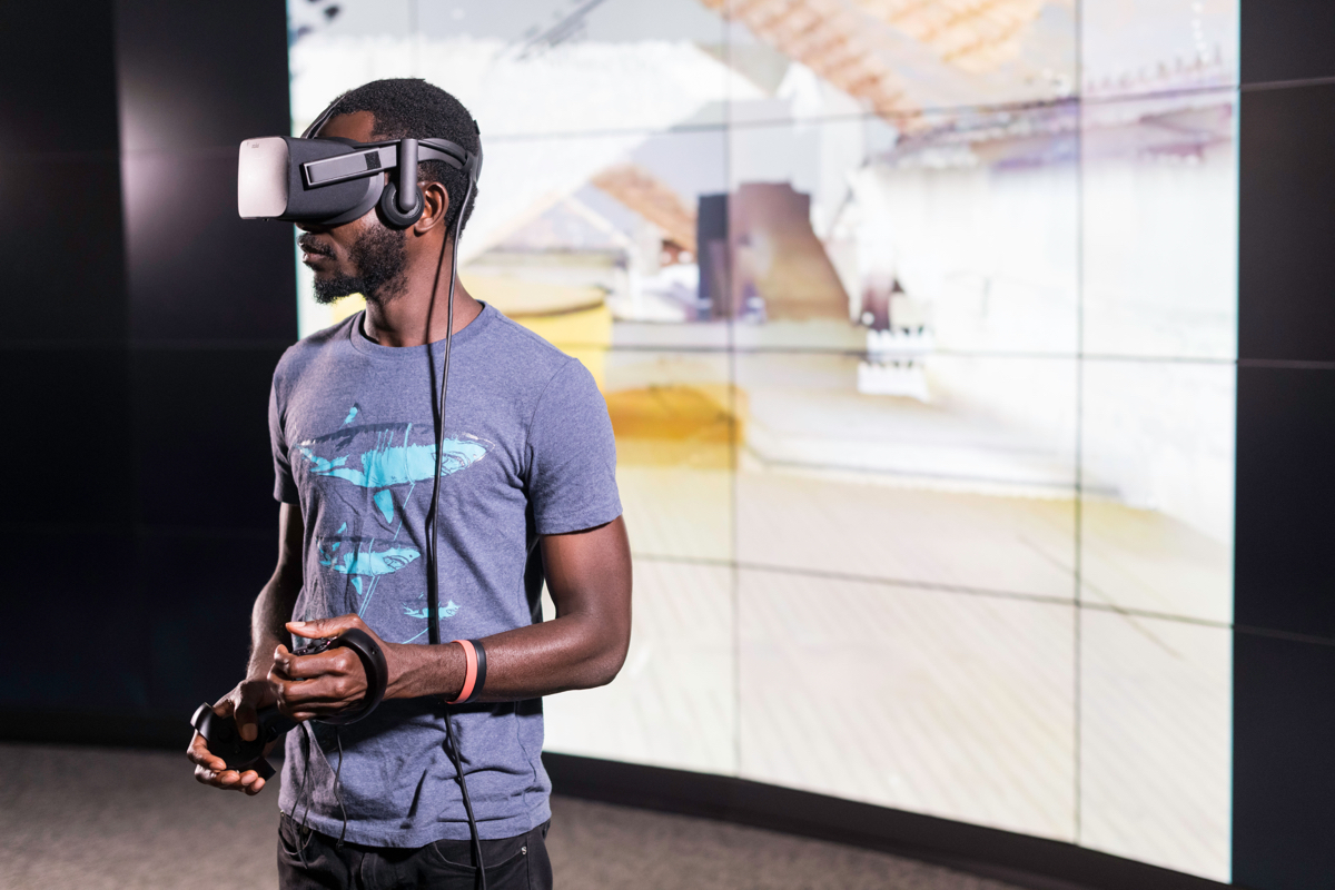 A University of Rochester student wearing a virtual reality headset stands in front of a large screen displaying graphics.