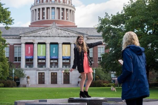 A young adult poses for a photo in front of the University of Rochester's Rush Rhees Library.