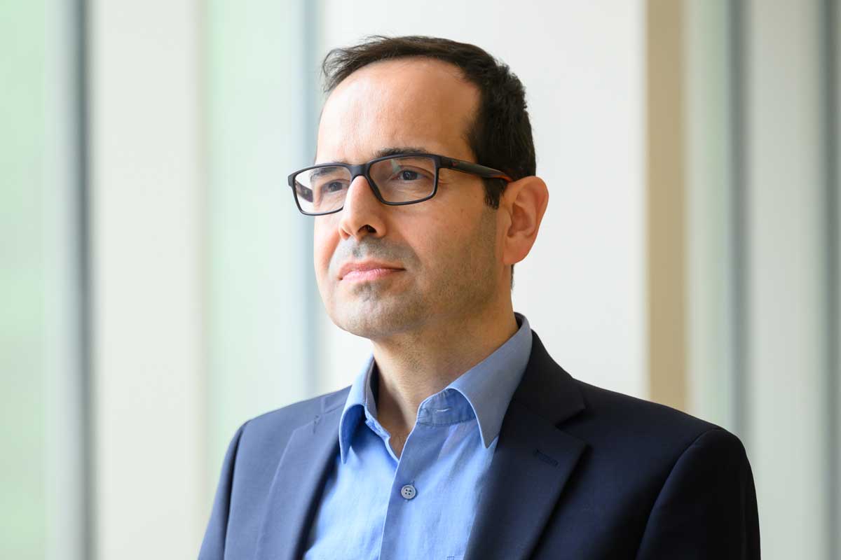Mujdat Cetin, the University of Rochester’s inaugural associate vice president and vice provost for artificial intelligence, stands near a window wearing glasses and a navy blazer.