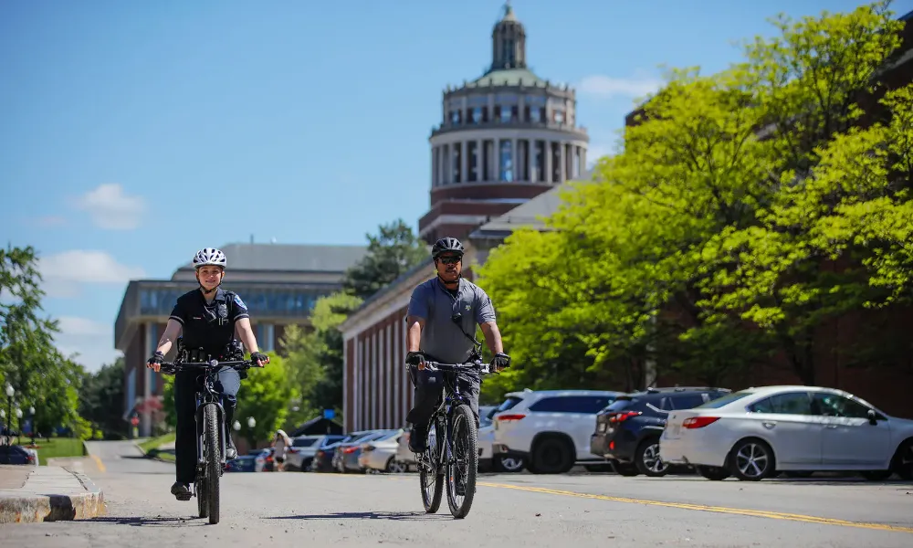 Two cyclists ride on a street in front of a University of Rochester building.