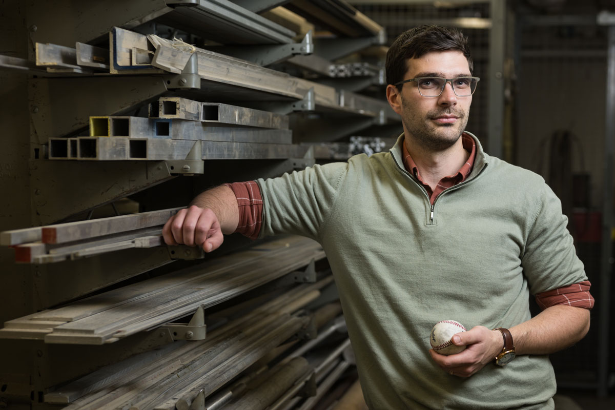 A man wearing glasses holds a baseball in a warehouse, representing baseball research at the University of Rochester.