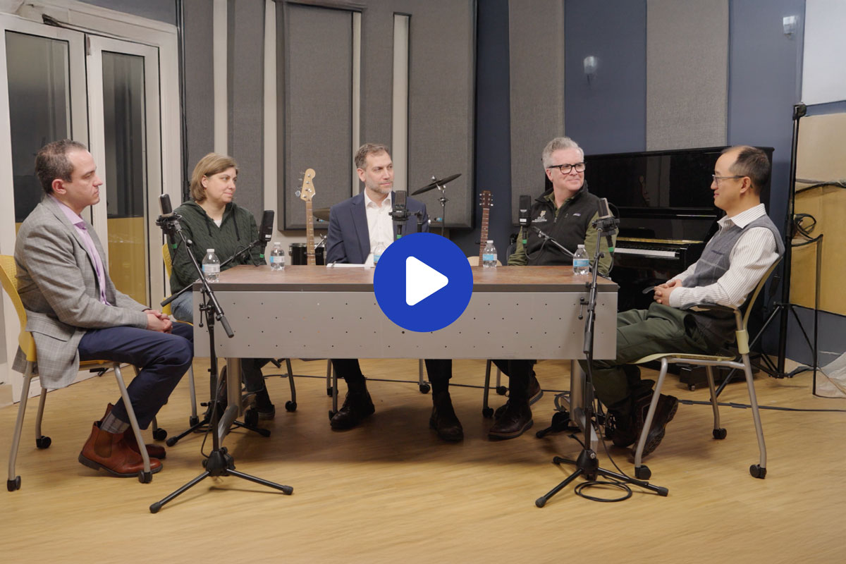 Four individuals seated at a table in a recording studio, engaged in discussion with audio equipment in the background.