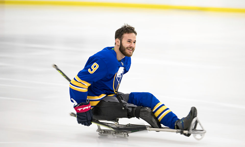 Becker, smiling and gliding across ice rink in Sabres uniform, mounted on hockey sled.