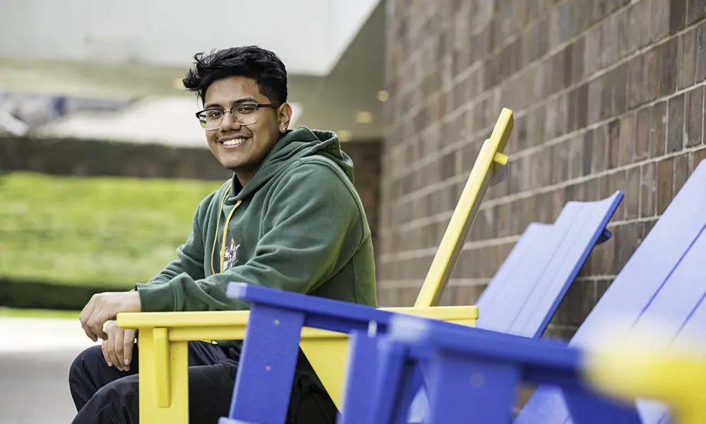 First-generation student Justin Pimentel seated outdoors in a yellow Adirondack chair and smiling at the camera.