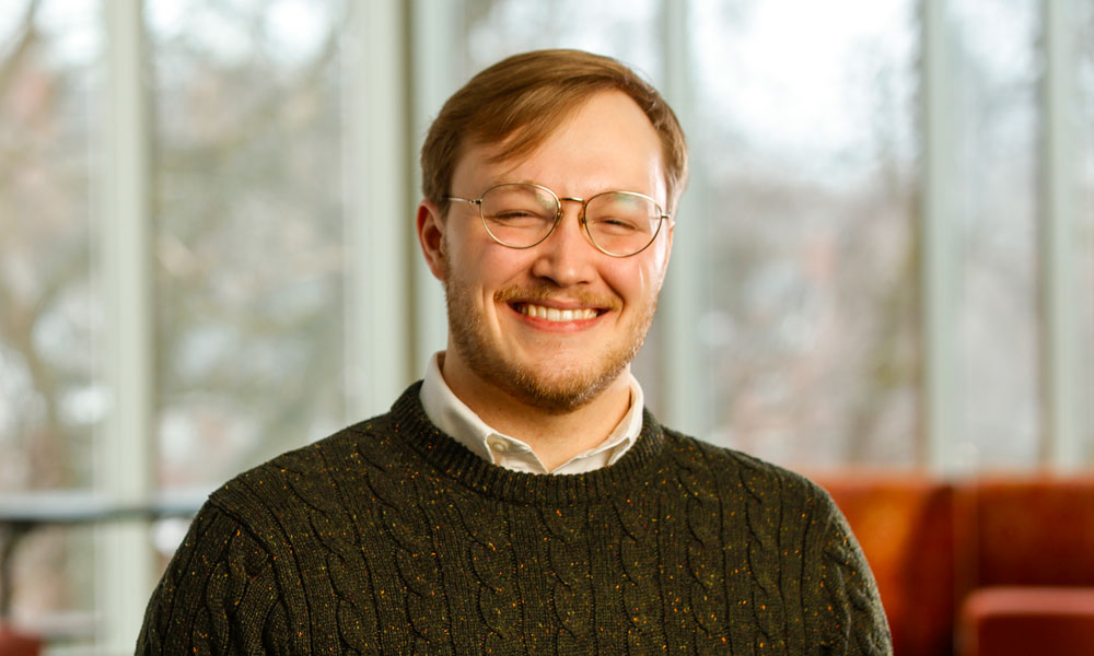 A smiling man with glasses and a sweater, Bryce Davis ’20, poses for the camera at the University of Rochester.