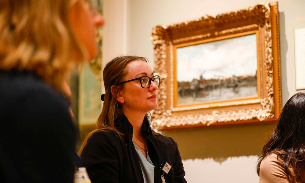 A woman wearing glasses and a black shirt observes a painting with a thoughtful expression.