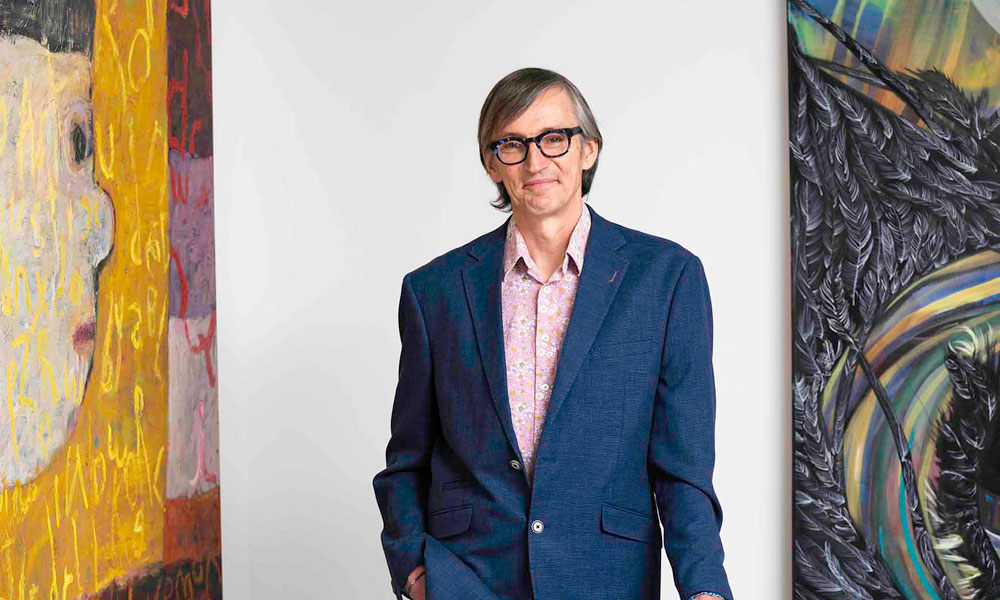 Stephen Dewhurst stands with his hand on a chair between two large paintings. .