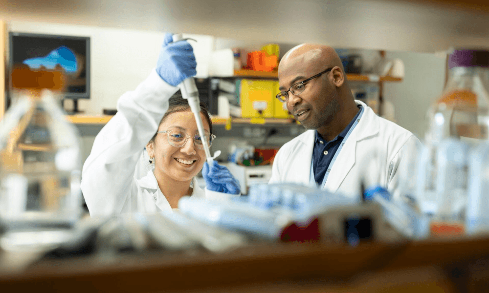 Two people wearing labcoats in a lab setting one holding a pipette while the other watches