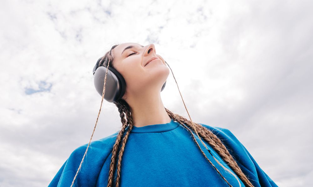 A young woman with pigtails wearing headphones and a blue sweatshirt is listening to music against the sky from a low wide angle.